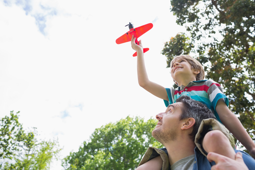 Low angle view of a boy with toy aeroplane sitting on fathers shoulders at the park